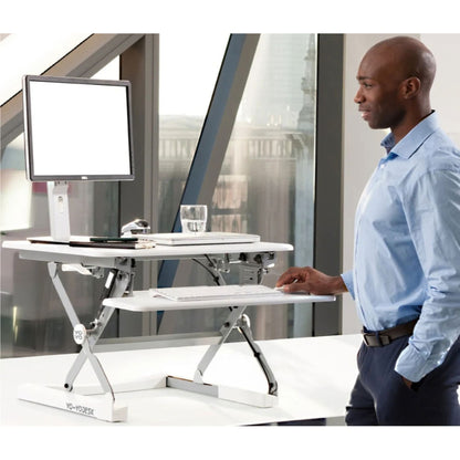 A man in a light blue dress shirt stands beside a white adjustable standing desk with a computer monitor, keyboard tray, and office accessories in a bright office setting.